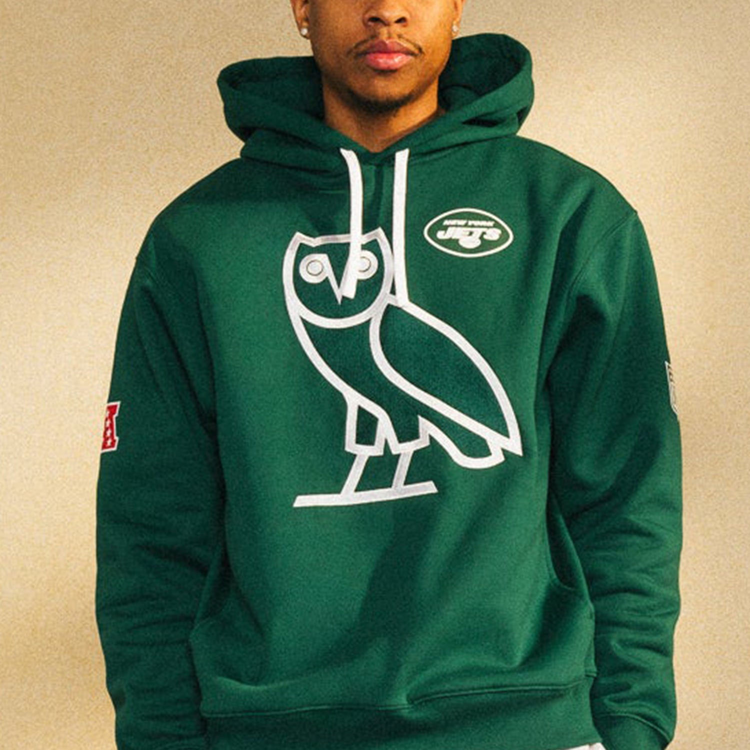 Man wearing a green New York Jets hoodie with an OVO owl logo in white on the front.